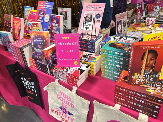A table with a pink table cloth displaying piles of paperback novels and tote bags with a sign saying ' PRICES - all books £5, 2 for £8, card payment only'